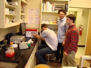 Cory Geiger loading samples into the flow cytometer as Stephen Woodall and John Collie look on with anticipation and excitement to come about the data!